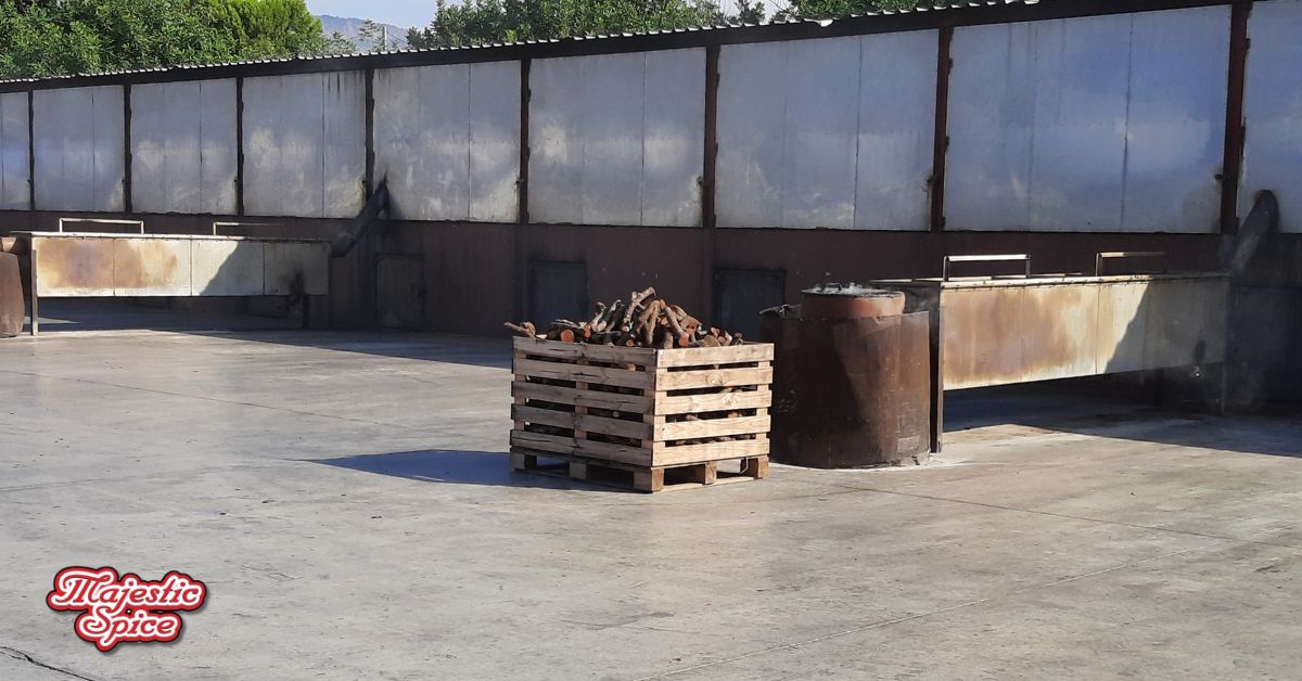 Outdoor area of a spice production facility showing a wooden crate filled with logs, positioned in front of large metal smoking or drying units, all under a clear blue sky. The logo 'Majestic Spice' is visible at the bottom left corner of the image.