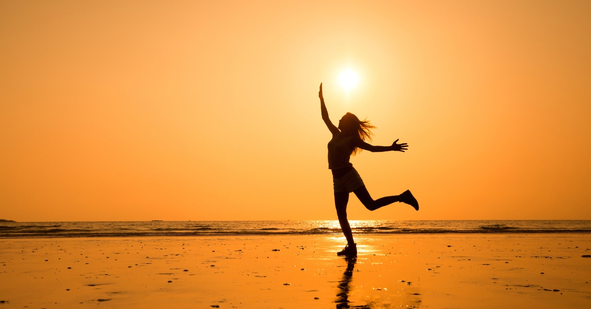 In this image we can see a lady standing in one foot with her arms in the air and her head a little bit towards the sky, like if she was dancing at the beach. The image is yellow with the sun up in her head and the sea at the background. She's at the shore and we can just see her shadow.