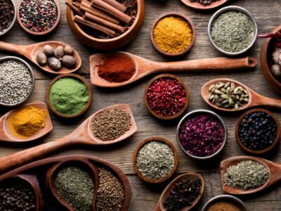 The image showcases an assortment of spices displayed in wooden bowls and spoons on a rustic wooden surface. The spices include various whole and ground forms such as cinnamon sticks, cardamom pods, black peppercorns, red chili peppers, turmeric powder, green powder, and dried herbs. The vibrant colors and textures of the spices are prominently featured, creating a visually appealing and aromatic display. In the bottom right corner, there is a logo with the text "Majestic Spice.
