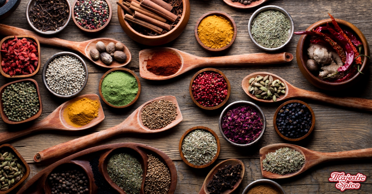 The image showcases an assortment of spices displayed in wooden bowls and spoons on a rustic wooden surface. The spices include various whole and ground forms such as cinnamon sticks, cardamom pods, black peppercorns, red chili peppers, turmeric powder, green powder, and dried herbs. The vibrant colors and textures of the spices are prominently featured, creating a visually appealing and aromatic display. In the bottom right corner, there is a logo with the text "Majestic Spice.
