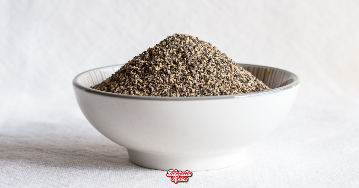 A white bowl filled with coarsely ground black pepper sits on a light-colored surface. The pepper granules are a mix of black and tan hues, showcasing the fresh and natural quality of the spice. The bowl is simple and elegant, with the "Majestic Spice" logo subtly displayed below.