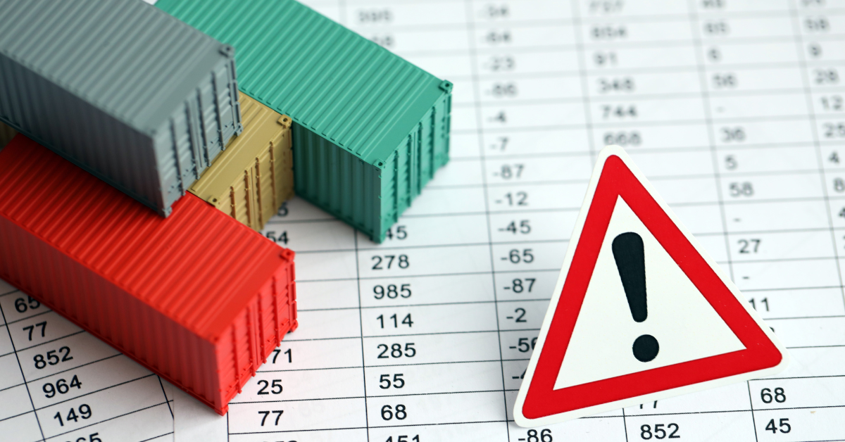 Stacked miniature shipping containers in various colors (red, teal, beige, gray) placed on a spreadsheet with numbers, alongside a triangular warning sign with an exclamation mark, symbolizing trade or logistics risk or disruption.