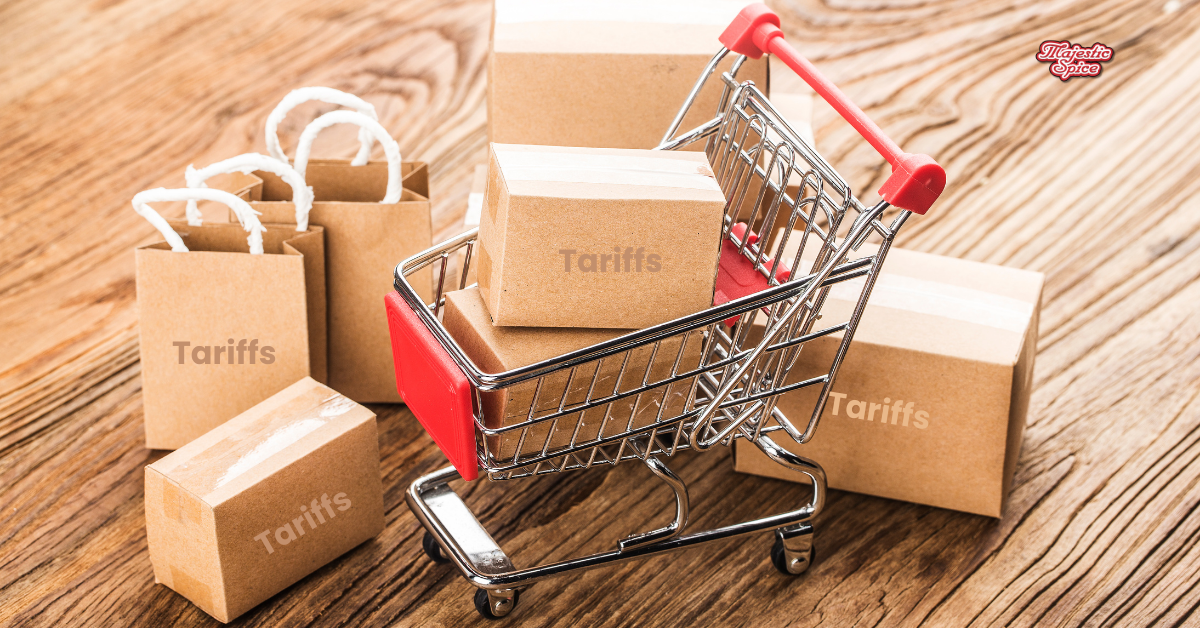 A miniature shopping cart filled with small cardboard boxes labeled "Tariffs," surrounded by more packages and paper bags with the same label, symbolizing the impact of tariffs on consumer goods. The Majestic Spice logo is visible in the top right corner.