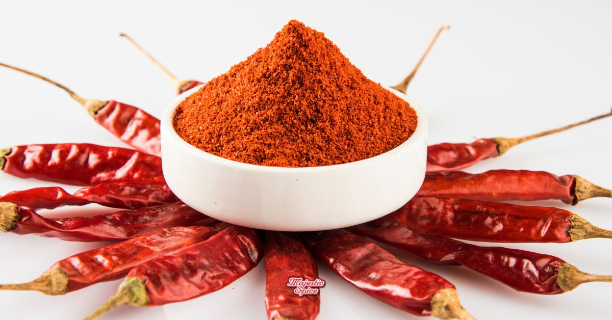 White bowl of red chili powder surrounded by whole dried red chilies on a white background.