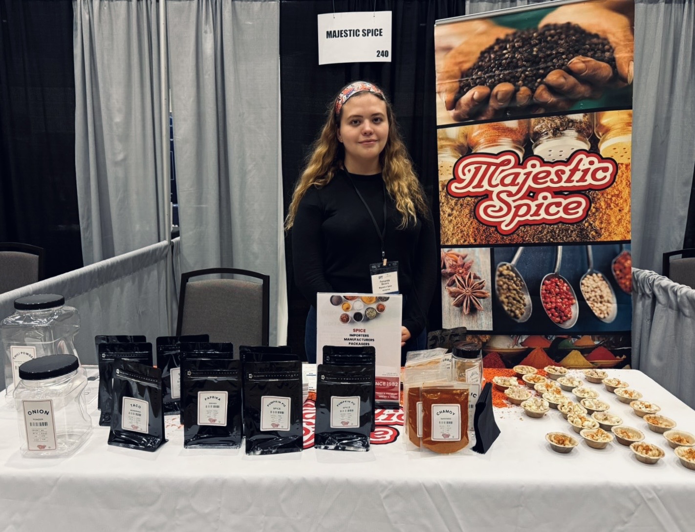 A young woman stands behind a Majestic Spice trade show booth. She is wearing a black top and name badge. The booth displays black sample pouches, seasoning jars, and small tasting cups arranged on a white table. Behind her is a large Majestic Spice banner featuring images of spices and a sign reading “MAJESTIC SPICE — 240.