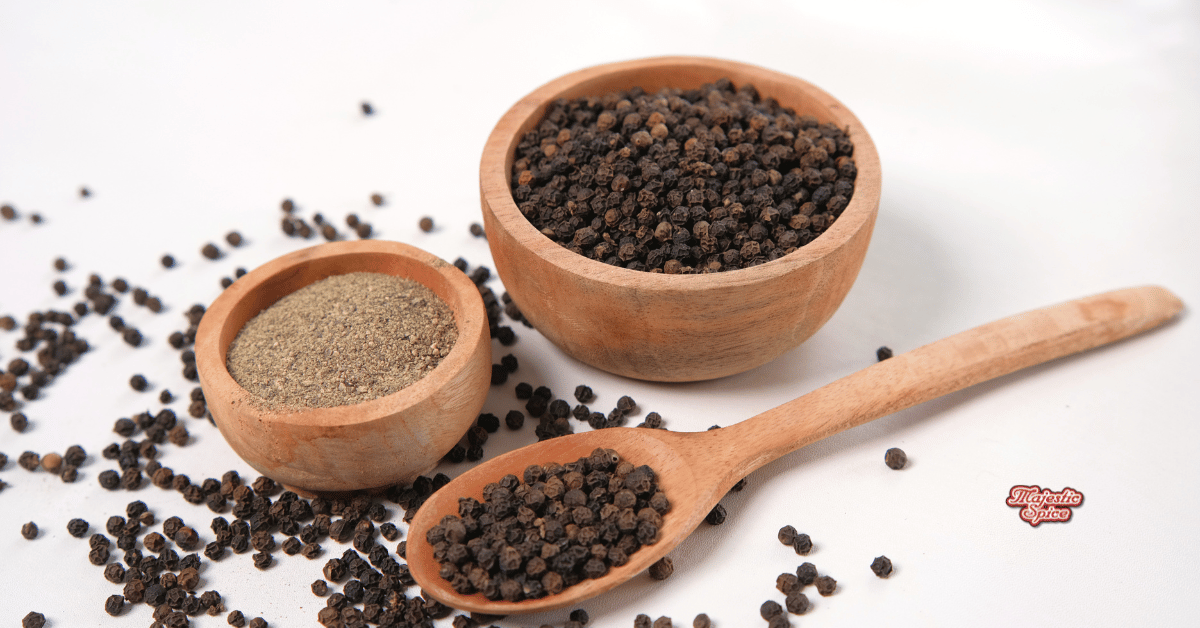 Whole and ground black pepper displayed in wooden bowls and a spoon on a white surface, with scattered peppercorns around. Majestic Spice logo visible in the bottom corner.