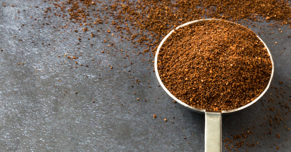 Close-up of a metal measuring cup filled with brown ground seasoning on a dark gray surface, with loose spice scattered around the cup.
